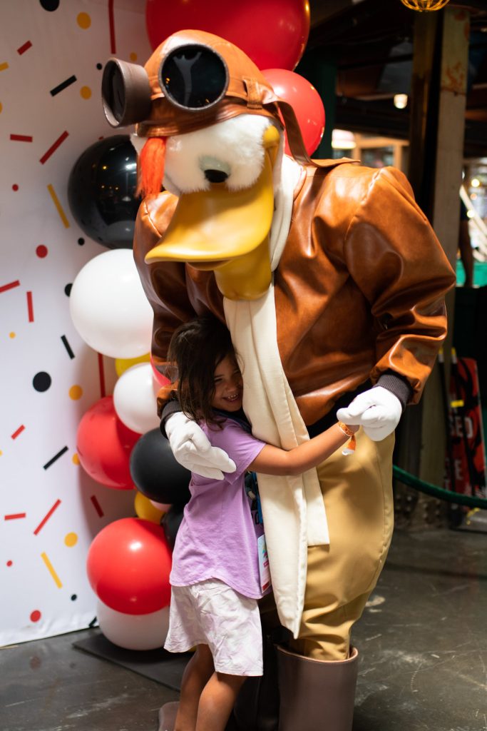 Launchpad McQuack from Tailspin hugging a little girl. The girl is wearing a purple tee shirt and white shorts. Launchpad is in his flight suit. He has aviator goggles on his head and is wearing a matching brown leather jacket and white scarf. The pair are standing in front of a confetti covered backdrop and red, white, and black balloons.