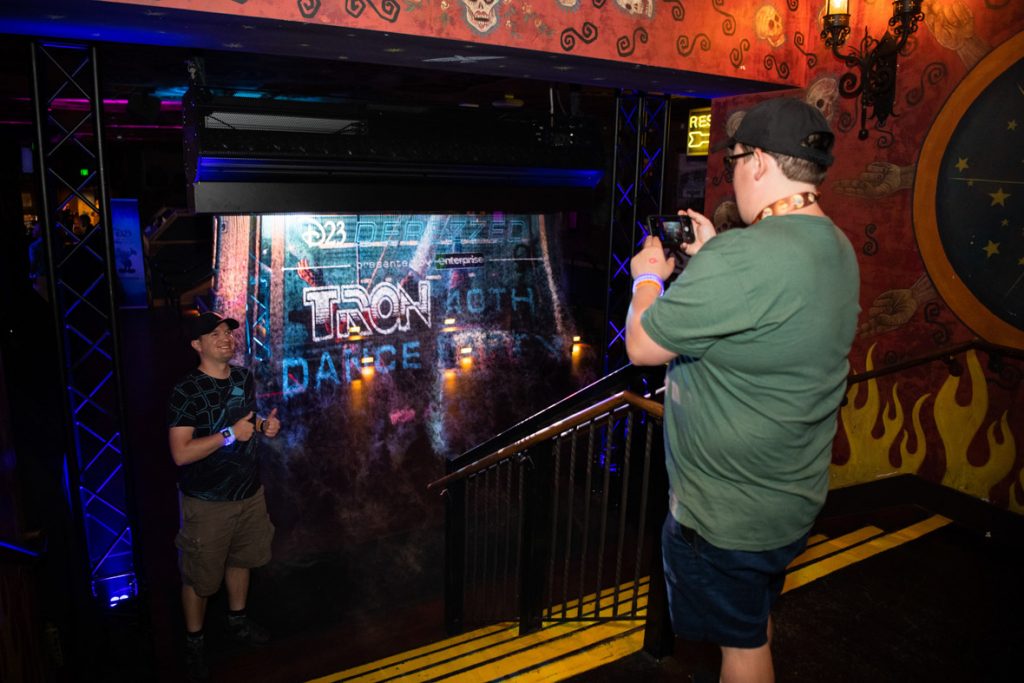 A fan takes a photo of another fan standing next to the mist screen. The fan taking the photo is wearing a black baseball hat, green T-shirt and black shorts holding their phone to take a photo positioned at the top of the stairs. At the bottom of the stairs is a fan standing to the left of the mist screen wearing a black baseball hat, black T-shirt and cargo shorts posing with two thumbs up. The mist screen features the D23 Derezzed Tron 40th Dance Party Logo in white and blue.