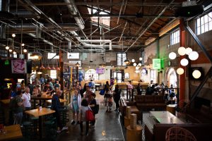 A full area photo of the event location, Punchbowl Social. Featured in the picture are fans mingling about. Lights are hung from the ceiling, and there is an industrial feel to the location. Giant Jenga games are featured in the lounge area, which includes tables and couches. Lots of daylight is brought in through the large open windows and doors around the venue. Retro video games are lined against one of the walls near high-top tables with green stools.
