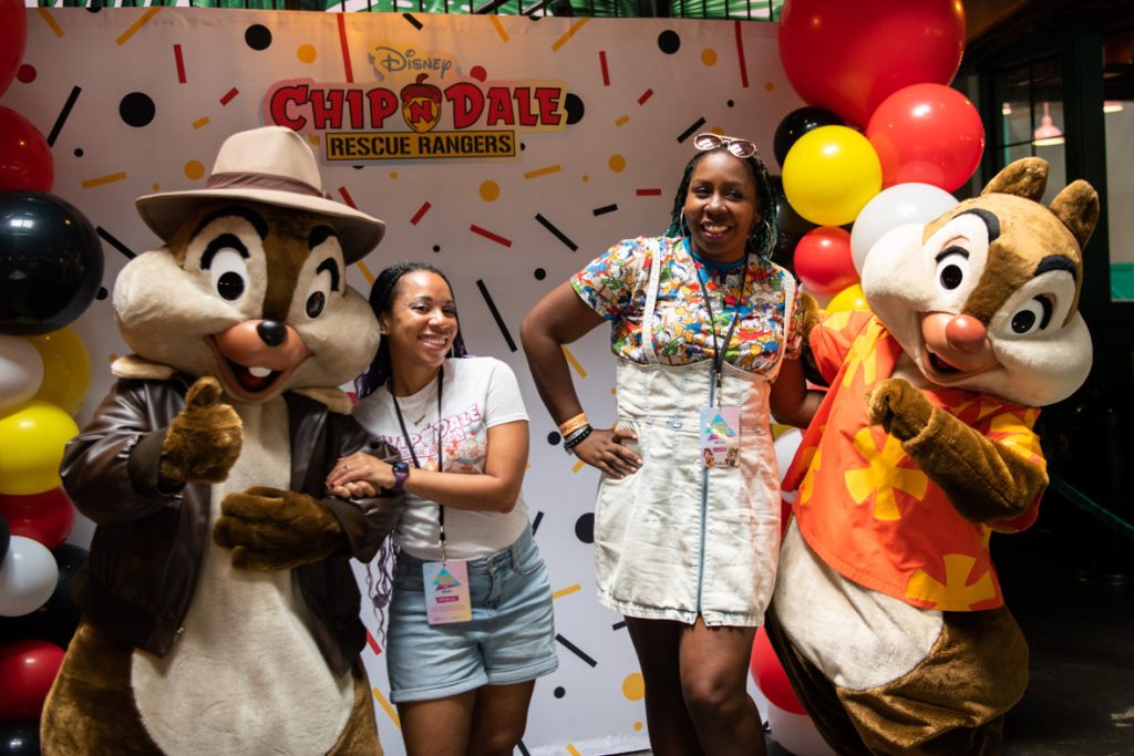 Two fans posing with Chip and Dale in their Rescue Rangers costumes. Chip is wearing a fedora style brown hat with a brown leather jacket. Dale is wearing a red Hawaiian style shirt with yellow flowers. Chip is on the far left holding their arm out for the fan next to him with his other hand in a thumbs up position. The fan has long dark hair with a white shirt with Chip and Dale Rescue Rangers in the center and denim shorts. The fan next has their arm around Dale with long dark hair, sunglasses on their head and hoop earrings. They are wearing a colorful printed shirt packed with DuckTales characters and a light denim overall dress. Dale has his arm around the fan and his other arm bent in front of him. All are smiling ear to ear in front of the photo backdrop that is white with red, yellow and black confetti shapes, there are red, white, yellow, and black balloon towers on either side and a Chip ’n Dale Rescue Rangers logo in the top center of the backdrop.