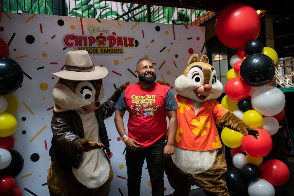 Fan smiling and posing with Chip and Dale Rescue Rangers. Chip on the left is pointing to the fan’s shirt with his arm on the fan’s shoulder. Chip is wearing a fedora style brown hat with a brown leather jacket. The fan is in the middle with a red shirt with grey sleeves featuring the Chip ’n Dale Rescue Rangers logo and characters in the center with black jeans. Dale has his arm on the fan on the right side with his other arm out. Dale is wearing a red Hawaiian style shirt with yellow flowers. They are all in front of the photo backdrop that is white with red, yellow and black confetti shapes, there are red, white, yellow, and black balloon towers on either side and a Chip ’n Dale Rescue Rangers logo in the top center of the backdrop.