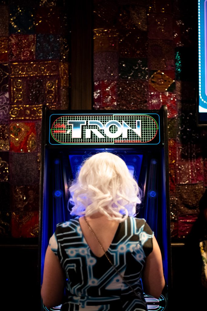 Back of fan with short platinum hair and black short sleeved dress with Tron-inspired blue and white lines. The fan is playing a retro cabinet Tron game. On the game, you can see the Tron logo on top of the cabinet in white letters with red and green grid lines behind it.