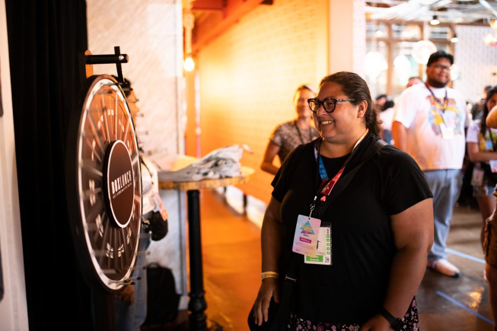 A fan with glasses, black shirt and floral skirt is smiling waiting to see what number she lands on at the BoxLunch Prize Wheel that is 5 feet tall, black, has a list of numbers one through 24 and BoxLunch’s logo featured in the middle. Behind the fan, multiple other fans are waiting for their turn to spin.