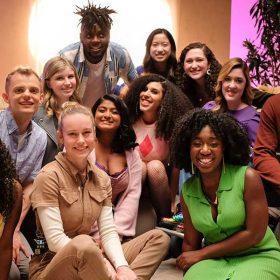 The Heroes and stars of Growing Up—Alex Crotty; Clare Della Valle and Isabel Lam; Amiri Nash; Athena Nair; Sofia Ongele; Emily Flores; Sage Grace Dolan-Sandrino; Gavin Arneson; Vanessa Aryee; and David Puma—gather around creator, executive producer, and director Brie Larson. Tan pillows and shapes are seen behind them, along with a leafy potted plant and bright light.