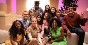 The Heroes and stars of Growing Up—Alex Crotty; Clare Della Valle and Isabel Lam; Amiri Nash; Athena Nair; Sofia Ongele; Emily Flores; Sage Grace Dolan-Sandrino; Gavin Arneson; Vanessa Aryee; and David Puma—gather around creator, executive producer, and director Brie Larson. Tan pillows and shapes are seen behind them, along with a leafy potted plant and bright light.