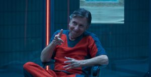 Actor Tim Roth as Emil Blonsky/Abomination sits in a chair and wears a bright orange jumpsuit and flexes his right arm like a finger gun. He sits in a clear prison cell that has no visible door.