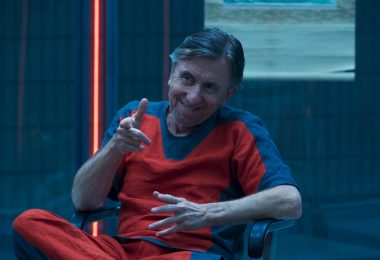Actor Tim Roth as Emil Blonsky/Abomination sits in a chair and wears a bright orange jumpsuit and flexes his right arm like a finger gun. He sits in a clear prison cell that has no visible door.