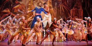 Actors Michael James Scott (Genie) and Michael Maliakel (Aladdin) take center stage with gold towers and riches in the background, and an ensemble of smiling dancers with batons before them in a showstopping scene from Aladdin on Broadway.