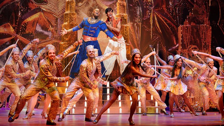 Actors Michael James Scott (Genie) and Michael Maliakel (Aladdin) take center stage with gold towers and riches in the background, and an ensemble of smiling dancers with batons before them in a showstopping scene from Aladdin on Broadway.