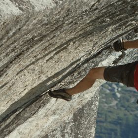 Alex Honnold free solos “Heaven” in Yosemite National Park. He is wearing a red T-shirt, black shirts, and climbing shoes, and is gripping onto a mountain.