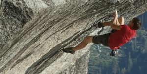 Alex Honnold free solos “Heaven” in Yosemite National Park. He is wearing a red T-shirt, black shirts, and climbing shoes, and is gripping onto a mountain.