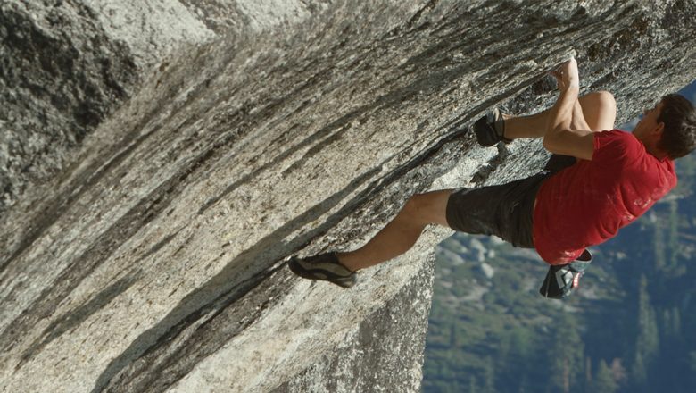 Alex Honnold free solos “Heaven” in Yosemite National Park. He is wearing a red T-shirt, black shirts, and climbing shoes, and is gripping onto a mountain.