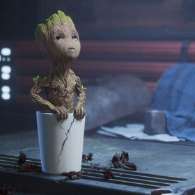 Baby Groot is planted in a cracked white pot and looking upward. The pot is on a workbench next to a wrench, and a blue tarp is in the background.