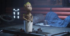 Baby Groot is planted in a cracked white pot and looking upward. The pot is on a workbench next to a wrench, and a blue tarp is in the background.