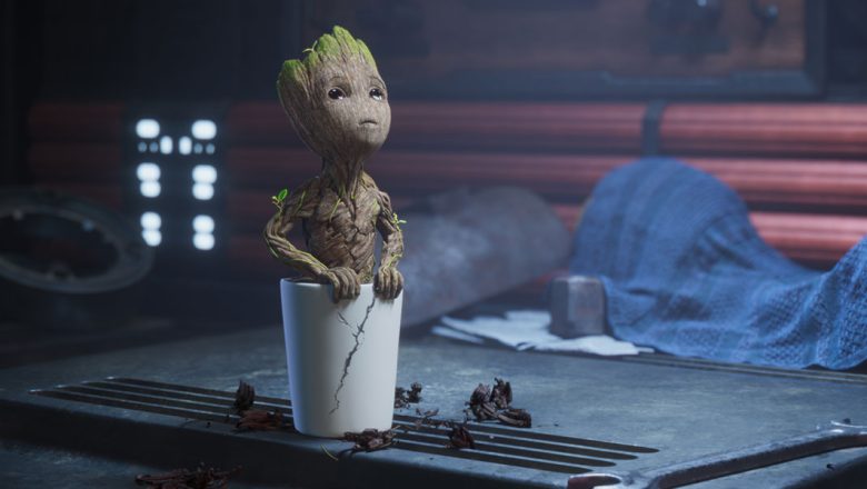 Baby Groot is planted in a cracked white pot and looking upward. The pot is on a workbench next to a wrench, and a blue tarp is in the background.