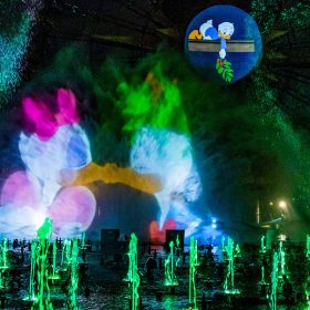 Dozens of water fountains are illuminated in green during “World of Color – Season of Light” at Disney California Adventure park. A central projection shows Daisy Duck and Donald Duck kissing with their eyes closed. Another smaller projection shows Dewey Duck lying face down on a mantle, holding mistletoe above Daisy and Donald.