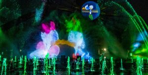 Dozens of water fountains are illuminated in green during “World of Color – Season of Light” at Disney California Adventure park. A central projection shows Daisy Duck and Donald Duck kissing with their eyes closed. Another smaller projection shows Dewey Duck lying face down on a mantle, holding mistletoe above Daisy and Donald.
