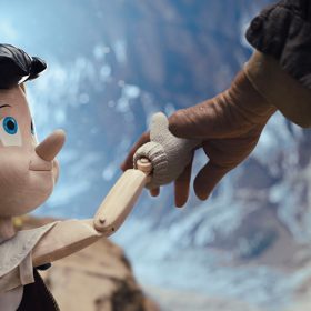 In a promotional still from Disney’s Pinocchio, the little wooden puppet is on the left, holding hands with and looking up at Geppetto (off-screen), on the right. Pinocchio is wearing a yellow felt hat with a blue stripe and a red feather, and a white collared shirt and dark vest.