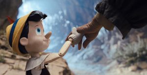 In a promotional still from Disney’s Pinocchio, the little wooden puppet is on the left, holding hands with and looking up at Geppetto (off-screen), on the right. Pinocchio is wearing a yellow felt hat with a blue stripe and a red feather, and a white collared shirt and dark vest.