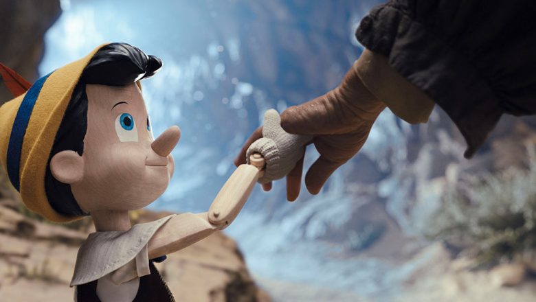 In a promotional still from Disney’s Pinocchio, the little wooden puppet is on the left, holding hands with and looking up at Geppetto (off-screen), on the right. Pinocchio is wearing a yellow felt hat with a blue stripe and a red feather, and a white collared shirt and dark vest.