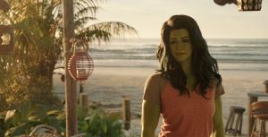 Actor Tatiana Maslany portraying Jennifer Walters/She-Hulk with CGI green skin and long green-black hair stands on a tropical beach with tall palm trees and bar tables behind her. She is wearing an orange tank top.