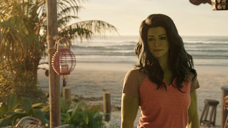 Actor Tatiana Maslany portraying Jennifer Walters/She-Hulk with CGI green skin and long green-black hair stands on a tropical beach with tall palm trees and bar tables behind her. She is wearing an orange tank top.