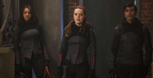 Actors Ashley Nicole Williams, Jessica Sutton, and Praneet Akilla hold robes in their hands and stand in front of a large steel door. Williams, Sutton, and Akilla wear all black outfits adorned with a small red stripe on their forearms.