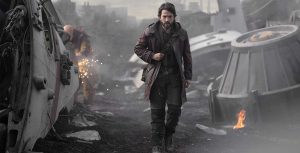 Diego Luna as Cassian Andor walks through wreckage of spaceships, eyes focused on something ahead. Behind him, a mechanic works on some scrap, sparks flying off of it. A piece of metal has caught fire next to some wreckage on the right.
