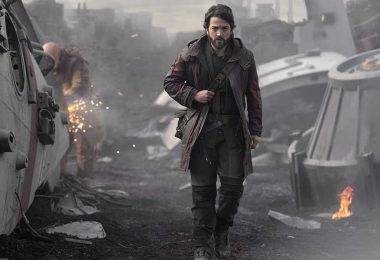 Diego Luna as Cassian Andor walks through wreckage of spaceships, eyes focused on something ahead. Behind him, a mechanic works on some scrap, sparks flying off of it. A piece of metal has caught fire next to some wreckage on the right.