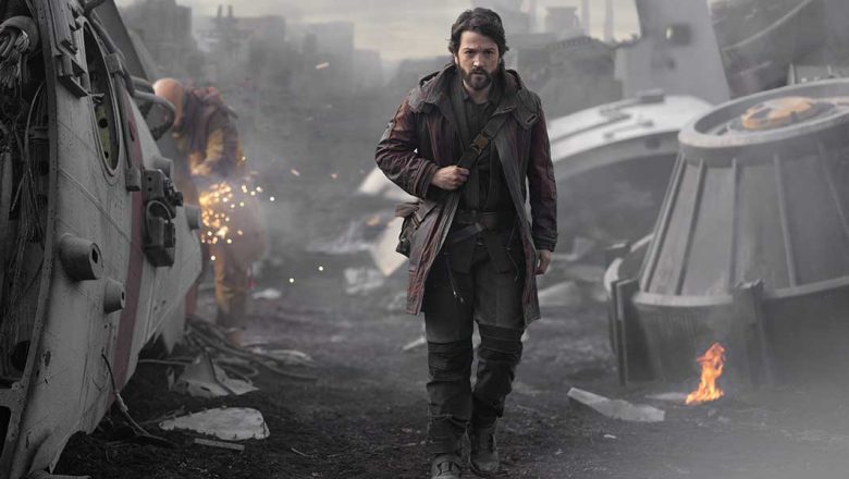 Diego Luna as Cassian Andor walks through wreckage of spaceships, eyes focused on something ahead. Behind him, a mechanic works on some scrap, sparks flying off of it. A piece of metal has caught fire next to some wreckage on the right.