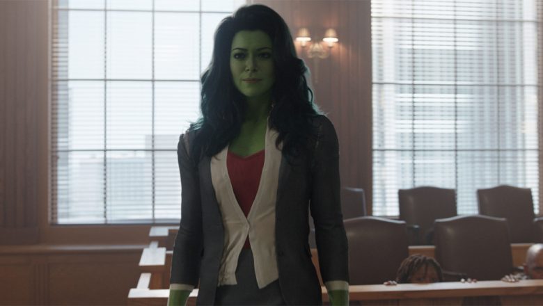 She-Hulk (Tatiana Maslany) stands in a courtroom. Her skin is bright green, and she has long wavy blue hair. She is wearing a red shirt with a white button up, and a gray blazer. The jury behind her is ducking behind the wood partition.