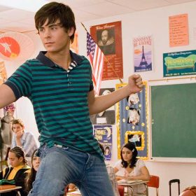 In a classroom, Zac Efron stands in his chair with one foot on his desk and flexes his biceps. He’s wearing a teal and navy striped polo and jeans. He is surrounded by seated co-stars including Ashley Tisdale, wearing a red bejeweled top and gold pants; Kacey Stroh, wearing a short-sleeve, heart-print hoodie and a lime green bangle; Lucas Grabeel, wearing a pink newsboy cap and a pink striped shirt; and Vanessa Hudgens, wearing a pink and white printed top and a white sweater. Corbin Bleu is standing behind Zac and wearing a green T-shirt that says, “I majored in VACATION,” plus a belt and jeans. Corbin is palming a basketball and looking at Zac.