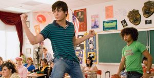 In a classroom, Zac Efron stands in his chair with one foot on his desk and flexes his biceps. He’s wearing a teal and navy striped polo and jeans. He is surrounded by seated co-stars including Ashley Tisdale, wearing a red bejeweled top and gold pants; Kacey Stroh, wearing a short-sleeve, heart-print hoodie and a lime green bangle; Lucas Grabeel, wearing a pink newsboy cap and a pink striped shirt; and Vanessa Hudgens, wearing a pink and white printed top and a white sweater. Corbin Bleu is standing behind Zac and wearing a green T-shirt that says, “I majored in VACATION,” plus a belt and jeans. Corbin is palming a basketball and looking at Zac.