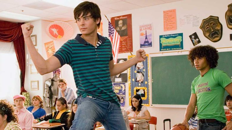 In a classroom, Zac Efron stands in his chair with one foot on his desk and flexes his biceps. He’s wearing a teal and navy striped polo and jeans. He is surrounded by seated co-stars including Ashley Tisdale, wearing a red bejeweled top and gold pants; Kacey Stroh, wearing a short-sleeve, heart-print hoodie and a lime green bangle; Lucas Grabeel, wearing a pink newsboy cap and a pink striped shirt; and Vanessa Hudgens, wearing a pink and white printed top and a white sweater. Corbin Bleu is standing behind Zac and wearing a green T-shirt that says, “I majored in VACATION,” plus a belt and jeans. Corbin is palming a basketball and looking at Zac.