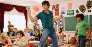 In a classroom, Zac Efron stands in his chair with one foot on his desk and flexes his biceps. He’s wearing a teal and navy striped polo and jeans. He is surrounded by seated co-stars including Ashley Tisdale, wearing a red bejeweled top and gold pants; Kacey Stroh, wearing a short-sleeve, heart-print hoodie and a lime green bangle; Lucas Grabeel, wearing a pink newsboy cap and a pink striped shirt; and Vanessa Hudgens, wearing a pink and white printed top and a white sweater. Corbin Bleu is standing behind Zac and wearing a green T-shirt that says, “I majored in VACATION,” plus a belt and jeans. Corbin is palming a basketball and looking at Zac.