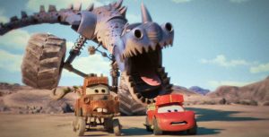 In an image from the Disney+ series Cars on the Road, the brown tow truck Mater is on the left and the red sports car Lightning McQueen is on the right. They’re being chased by some sort of purple dinosaur-car hybrid with large wheels through a desert landscape.