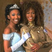 In a promotional still from 1997’s Wonderful World of Disney film Rodgers & Hammerstein’s Cinderella, Brandy is standing on the left in an iconic Cinderella-style light blue ballgown, white gloves, and a tiara, and she has her hand on Whitney Houston’s arm, who’s standing on the right in her golden Fairy Godmother costume, covered in jewels with a high collar.