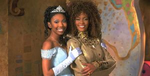 In a promotional still from 1997’s Wonderful World of Disney film Rodgers & Hammerstein’s Cinderella, Brandy is standing on the left in an iconic Cinderella-style light blue ballgown, white gloves, and a tiara, and she has her hand on Whitney Houston’s arm, who’s standing on the right in her golden Fairy Godmother costume, covered in jewels with a high collar.