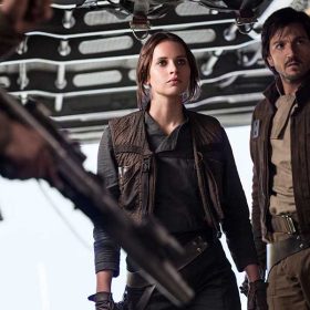 In a still from Rogue One: A Star Wars Story, Jyn Erso (Felicity Jones) and Cassian Andor (Diego Luna) are standing together near a ramp of a starship leading to outside, looking at several people in front of them holding weapons. They are wearing dark-colored clothing and both are wearing holstered weapons.