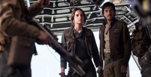 In a still from Rogue One: A Star Wars Story, Jyn Erso (Felicity Jones) and Cassian Andor (Diego Luna) are standing together near a ramp of a starship leading to outside, looking at several people in front of them holding weapons. They are wearing dark-colored clothing and both are wearing holstered weapons.