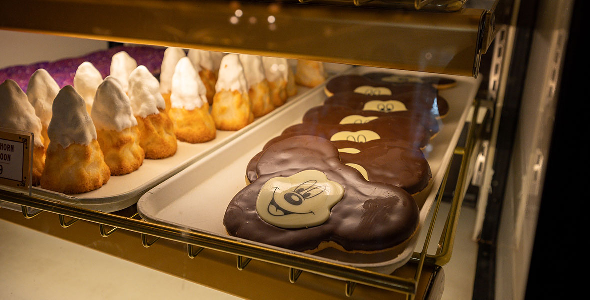 The Mickey-shaped cookies are covered in chocolate with a white cutout of Mickey Mouse’s face. The cookies are stacked on a metal tray inside of a bakery display next to other pastries.