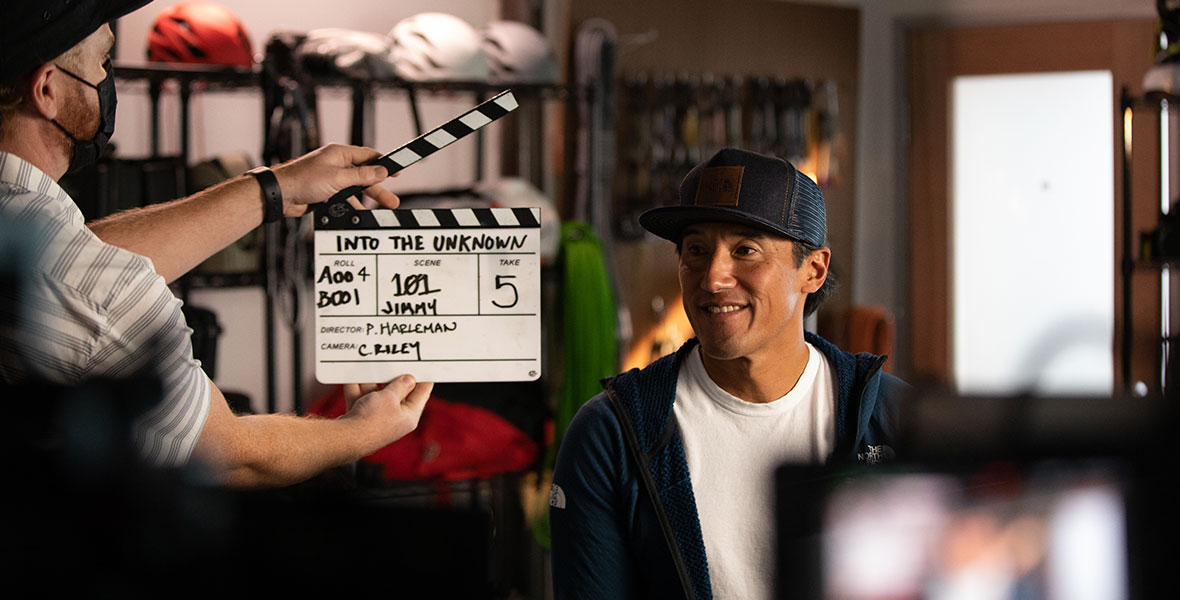Filmmaker and climber Jimmy Chin films an interview for Edge of the Unknown with Jimmy Chin. He is wearing a navy blue baseball cap with a brown leather patch, a white T-shirt, and navy blue Northface hoodie. He is looking to his right at a crew member, who is holding a black and white slate for scene 101, take 5.
