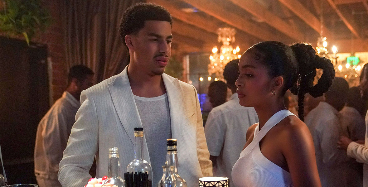Actors Marcus Scribner and Yara Shahidi stand at a small bar top covered with bottles of alcohol