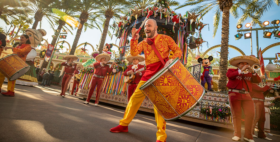 A drummer smiles wide as he bangs on his drum during the “Disney ¡Viva Navidad!” street party at Disney California Adventure park. He is wearing a patterned gold and red outfit, which matches his drum. He stands next to two other drummers, wearing similar outfits, and six Mariachis, wearing red jackets, red pants, white shirts, and hats with gold trim. Behind them are Mickey Mouse and Minnie Mouse, wearing rainbow fiesta attire. They are surrounded by palm trees near Paradise Gardens Park.