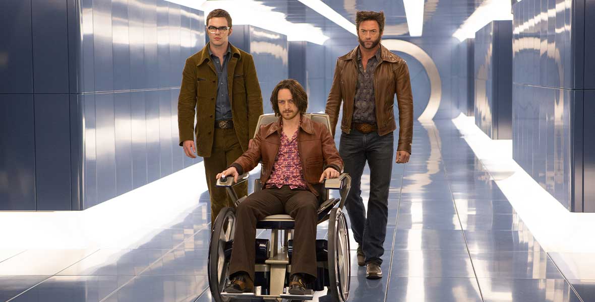 Actors Nicholas Hoult and Hugh Jackman walk down a hallway with dark blue tiles covering the walls and floors. Actor James McAvoy sits in a motorized wheelchair between Hoult and Jackman
