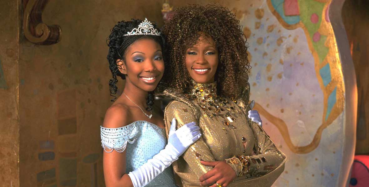 Actors and musical artists Brandy and Whitney Houston embrace while portraying Cinderella and Fairy Godmother, respectively. Brandy wears a light blue ball gown off her shoulders, matching gloves, and a silver tiara. Houston wears a gold dress with a high neckline and ornate beading.