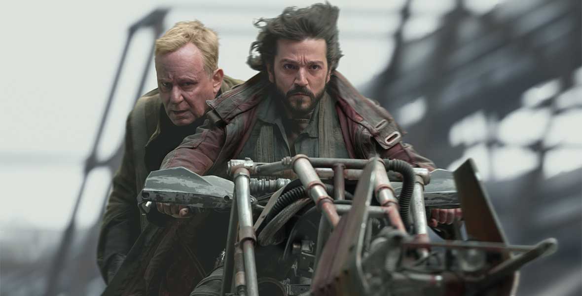 In a scene from Andor, Luthen Rael, played by Stellan Skarsgård, is riding a speeder bike. He is seated behind Cassian Andor, played by Diego Luna, who is steering the bike. The background is blurred and their hair is windblown, implying they are traveling at high speeds.