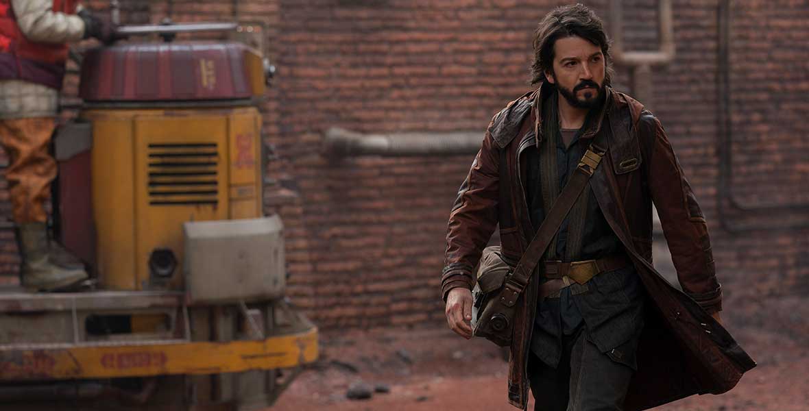 Diego Luna as Cassian Andor walks through a street, a brick building behind him. On the left of the image, a person driving some kind of yellow and red transportation vehicle