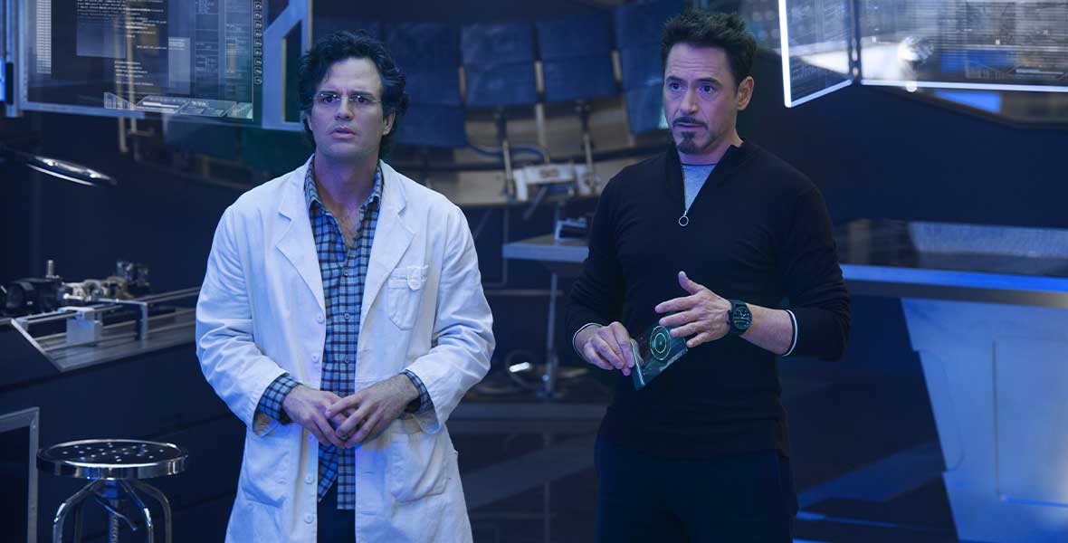 Tony Stark (Robert Downey Jr.) stands to the left of Bruce Banner (Mark Ruffalo). Banner is wearing a blue plaid shirt, black pants, and a white lab coat. He is wearing frameless glasses and has black wavy hair. Stark has facial hair and a black wristwatch and is holding a tablet. He is wearing a black zip top and black pants.