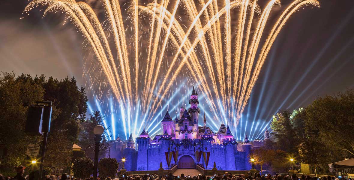 A time-lapse photo of the Believe... in Holiday Magic fireworks at Disneyland park, with plumes of white sparkling fireworks shooting up from behind Sleeping Beauty’s Castle, as well as columns of light. Many people are standing in front of the castle watching the display. The castle itself it bathed in a purple glow.